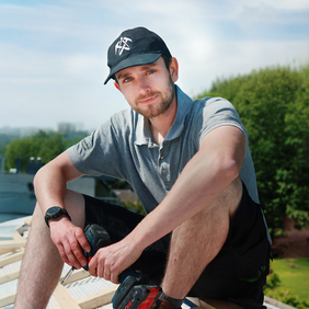 Porträt eines jungen Mannes im Arbeitsoutfit mit grauem Poloshirt, schwarzer Kappe und Armbanduhr. Er sitzt draußen auf einer Holzkonstruktion auf einem Dach, hält einen Akkuschrauber in der Hand und blickt direkt in die Kamera. Im Hintergrund sind Bäume und ein hellblauer Himmel zu sehen.
