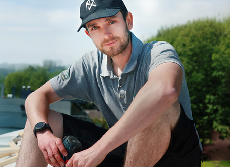 Porträt eines jungen Mannes im Arbeitsoutfit mit grauem Poloshirt, schwarzer Kappe und Armbanduhr. Er sitzt draußen auf einer Holzkonstruktion auf einem Dach, hält einen Akkuschrauber in der Hand und blickt direkt in die Kamera. Im Hintergrund sind Bäume und ein hellblauer Himmel zu sehen.